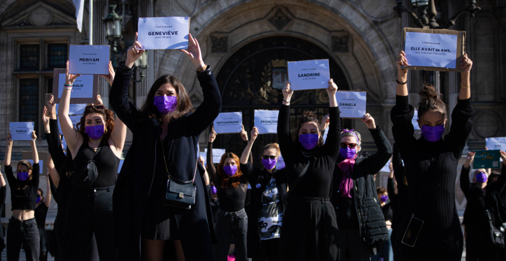 Des femmes habillées de noir et portant des masques violets tiennent à bout de bras des papiers avec le prénom de victimes de féminicides